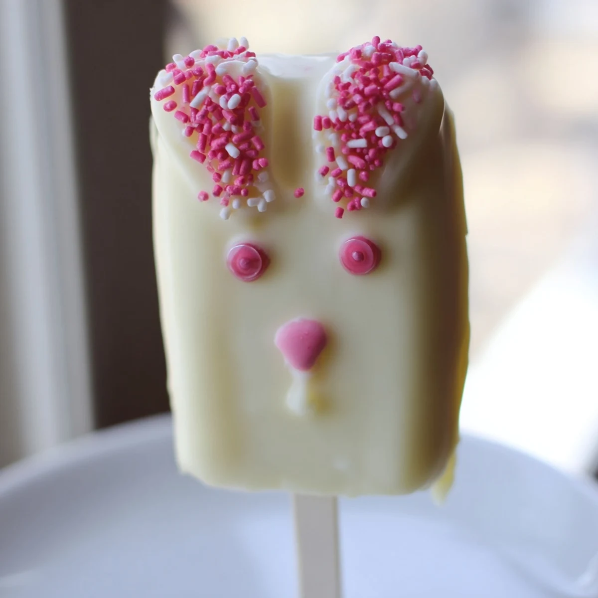 Creamy white chocolate bunny cakesicles decorated with pink ears and colorful sprinkles