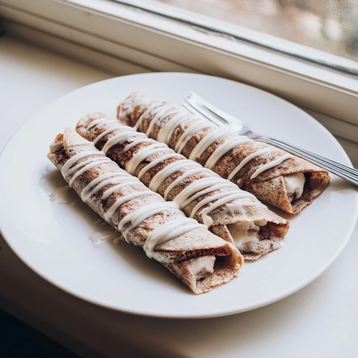 Golden cinnamon roll protein crepes stacked on a plate topped with powdered sugar glaze