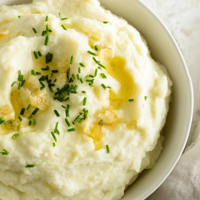 Silky smooth Roasted Garlic Mashed Cauliflower topped with chopped chives, next to roasted chicken on a dinner plate. Perfect gluten-free, low-carb holiday side.